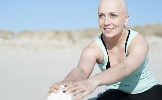 Mujer sin cabello, paciente oncológica, estirando en la playa con ropa deportiva